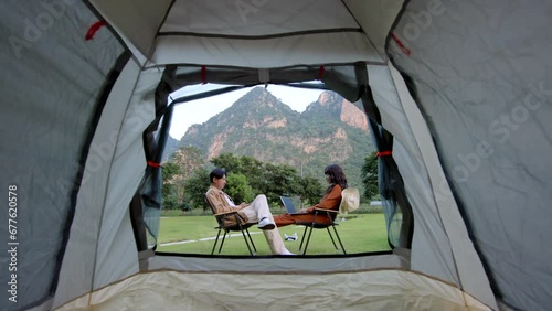 Modern people use technology connection in travel sitting in camp chairs near a tent. Handsome asian man showing contents on phone to his wife.