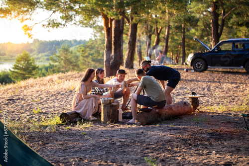 A company of friends of girls and guys is resting in the summer in a picturesque place in the forest on the banks of the river against the backdrop of sunset. Picnic in nature, tourism. Celebrating a