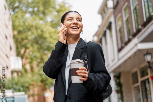 Stressless young businesswoman on the phone with a cheerful expression, in the street, holding a thermal cup of coffee