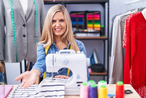 Blonde woman dressmaker designer using sew machine smiling cheerful offering palm hand giving assistance and acceptance.