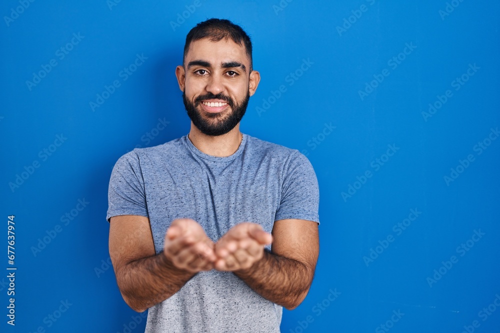 Naklejka premium Middle east man with beard standing over blue background smiling with hands palms together receiving or giving gesture. hold and protection