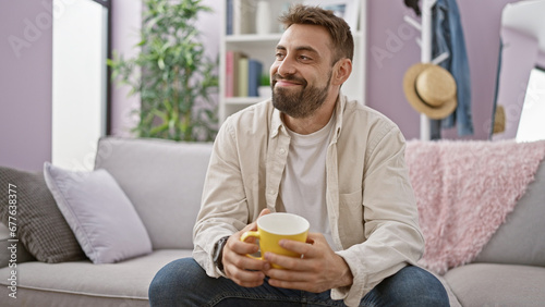 Joyful morning brew, young, bearded hispanic man radiates confidence and happiness, smiling, holding a coffee cup and resting in comfort on the sofa at his stylish apartment.