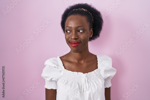 African woman with curly hair standing over pink background smiling looking to the side and staring away thinking.