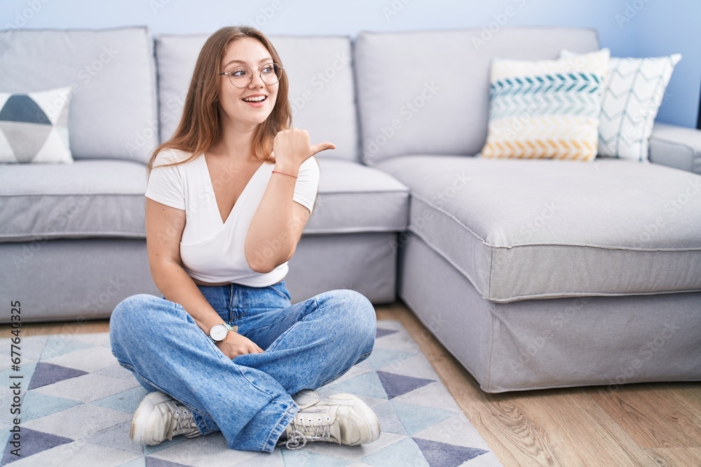 Young caucasian woman sitting on the floor at the living room smiling with happy face looking and pointing to the side with thumb up. Young caucasian woman sitting on the floor at the living room smiling with happy face looking and pointing to the side with thumb up.