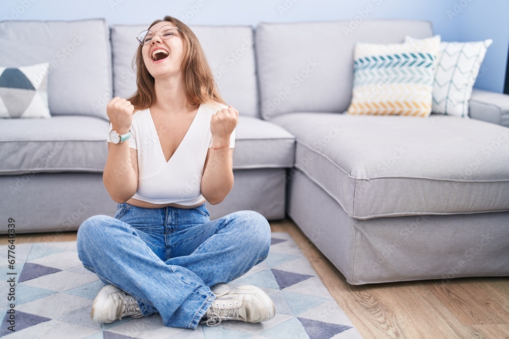 Young caucasian woman sitting on the floor at the living room celebrating surprised and amazed for success with arms raised and eyes closed. winner concept. Young caucasian woman sitting on the floor at the living room celebrating surprised and amazed for success with arms raised and eyes closed. winner concept.