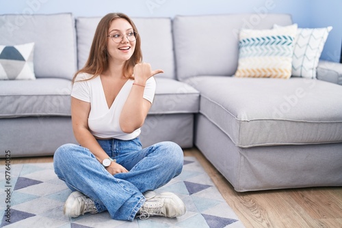 Young caucasian woman sitting on the floor at the living room smiling with happy face looking and pointing to the side with thumb up. © Krakenimages.com