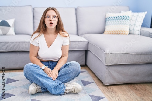 Photos Young caucasian woman sitting on the floor at the living room afraid and shocked with surprise and amazed expression, fear and excited face