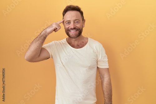 Middle age man with beard standing over yellow background smiling pointing to head with one finger, great idea or thought, good memory