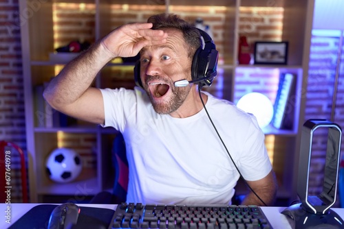 Middle age man with beard playing video games wearing headphones very happy and smiling looking far away with hand over head. searching concept. photo