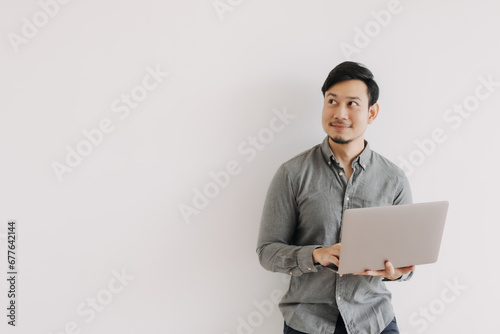 Asian man with beard wear grey shirt typing keyboard working on notebook and holding laptop, happy smiling face looking at empty space and standing isolated over white background wall.