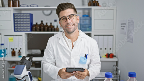 Handsome young hispanic man  confident smiling scientist  comfortably working with touchpad at his high-tech lab  advancing in the world of science and medicine