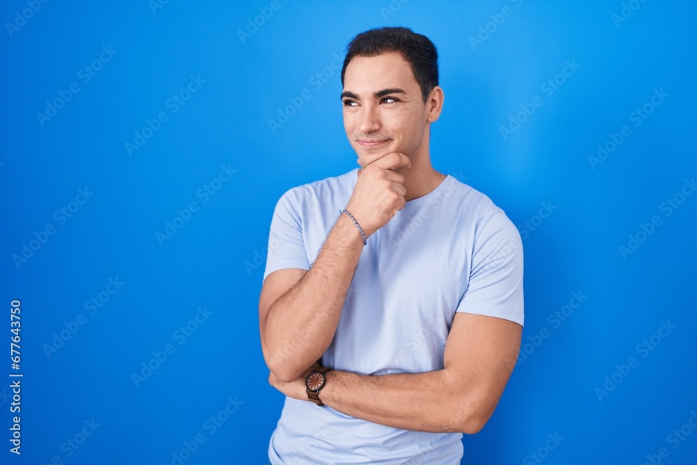 Young hispanic man standing over blue background looking confident at the camera smiling with crossed arms and hand raised on chin. thinking positive. Young hispanic man standing over blue background looking confident at the camera smiling with crossed arms and hand raised on chin. thinking positive.
