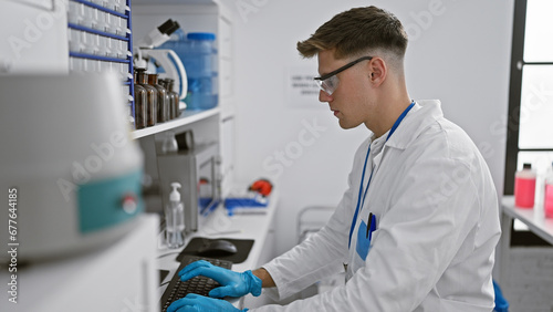 Attractive young caucasian male scientist totally engrossed in typing on computer amidst test tubes and microscopes  indoor laboratory work