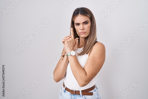 Hispanic young woman standing over white background holding symbolic gun with hand gesture, playing killing shooting weapons, angry face