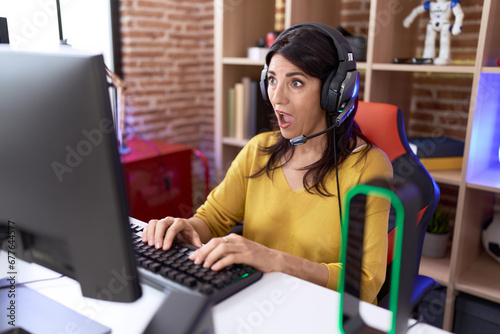 Middle age hispanic woman playing video games using headphones scared and amazed with open mouth for surprise, disbelief face