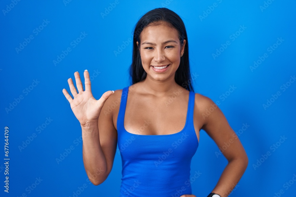 Hispanic woman standing over blue background showing and pointing up with fingers number five while smiling confident and happy. Hispanic woman standing over blue background showing and pointing up with fingers number five while smiling confident and happy.