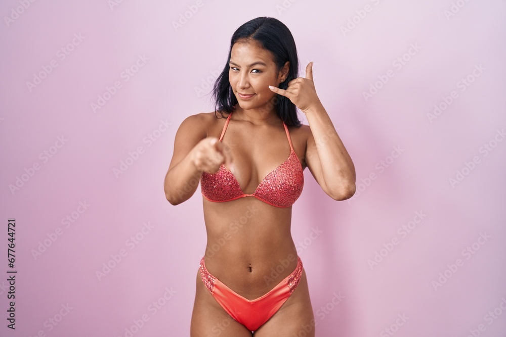 Naklejka premium Hispanic woman wearing bikini smiling doing talking on the telephone gesture and pointing to you. call me.