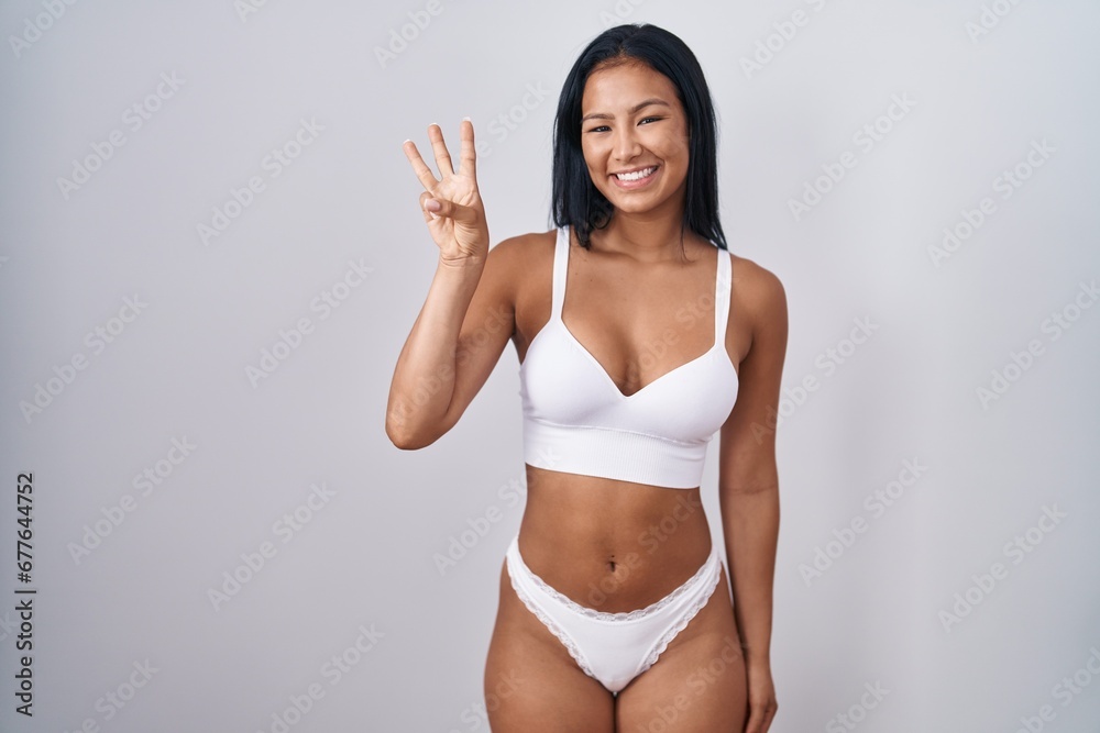 Fototapeta premium Hispanic woman wearing lingerie showing and pointing up with fingers number three while smiling confident and happy.