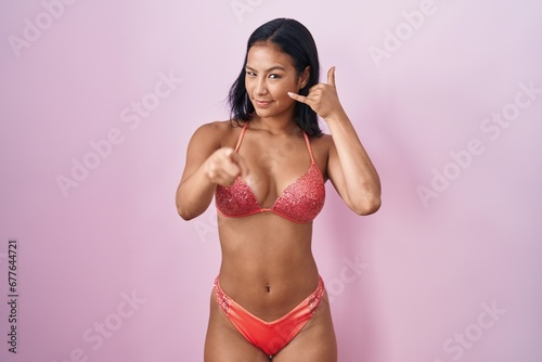 Hispanic woman wearing bikini smiling doing talking on the telephone gesture and pointing to you. call me.