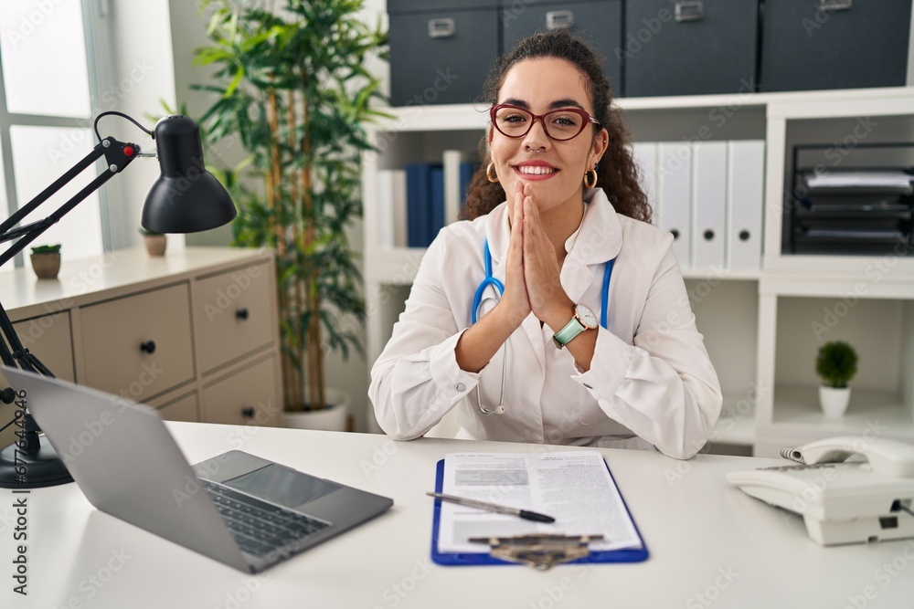 Young hispanic woman wearing doctor uniform and stethoscope praying with hands together asking for forgiveness smiling confident. Young hispanic woman wearing doctor uniform and stethoscope praying with hands together asking for forgiveness smiling confident.