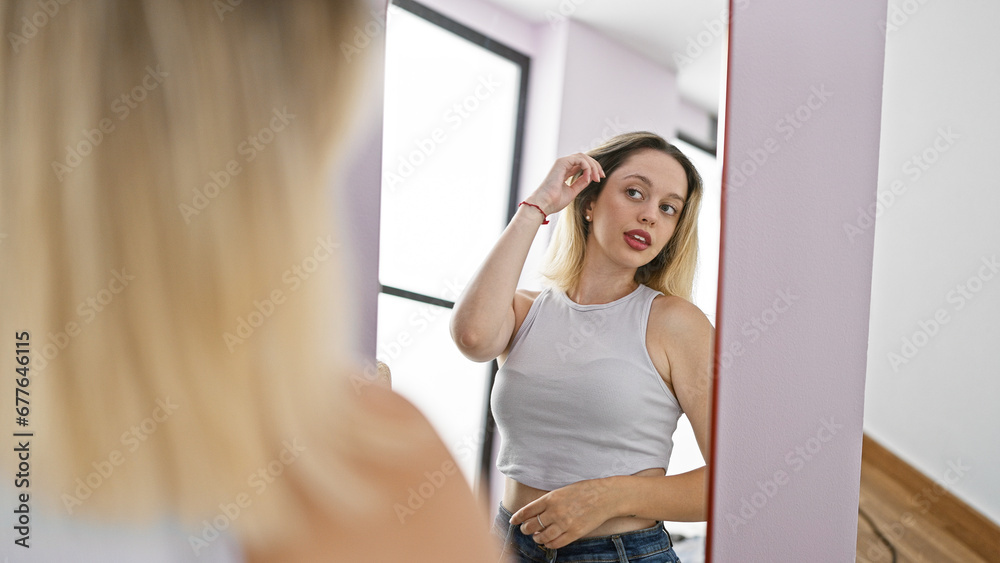 Young blonde woman combing hair with hands looking on mirror at home