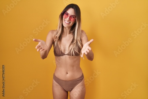 Young hispanic woman wearing bikini over yellow background smiling cheerful offering hands giving assistance and acceptance.