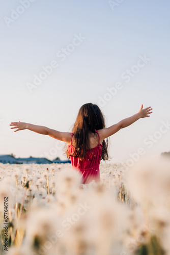 Little girl standing in white dandelions meadow with arms wide open. Female child enjoying summer sunny day in nature.