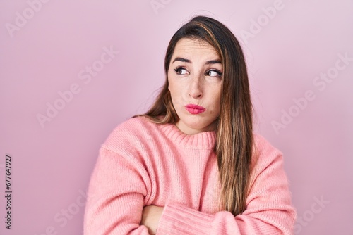 Young hispanic woman standing over pink background looking to the side with arms crossed convinced and confident