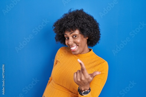 Black woman with curly hair standing over blue background beckoning come here gesture with hand inviting welcoming happy and smiling © Krakenimages.com