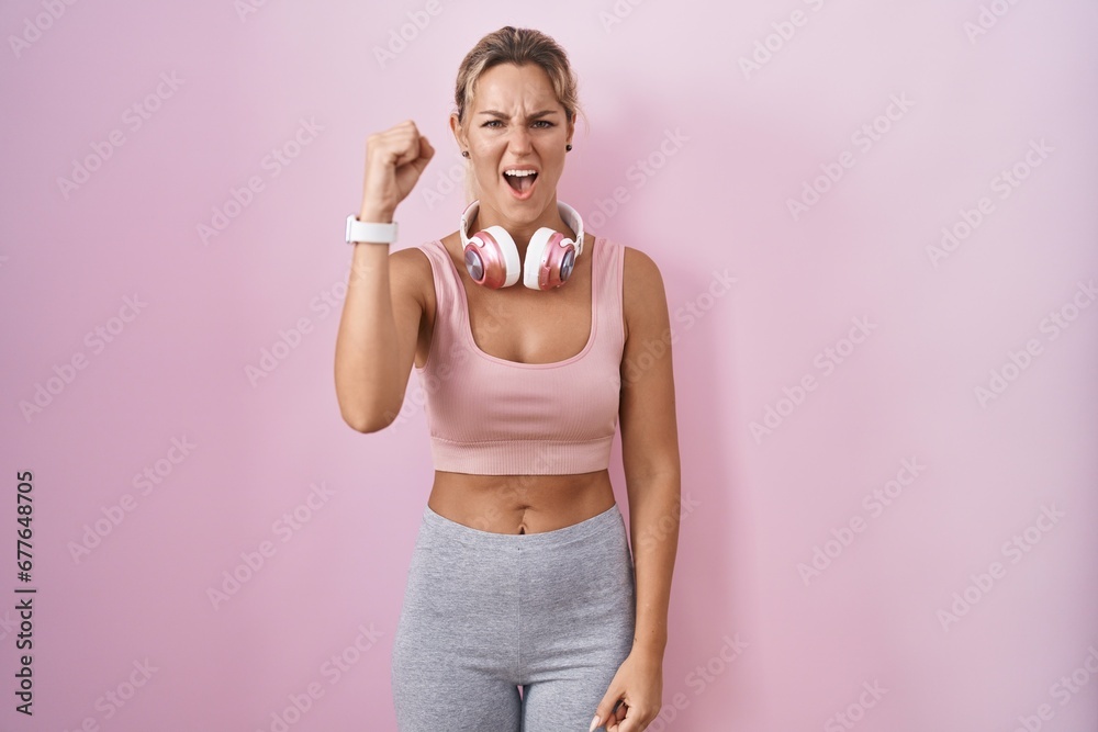 Young blonde woman wearing sportswear and headphones angry and mad raising fist frustrated and furious while shouting with anger. rage and aggressive concept.