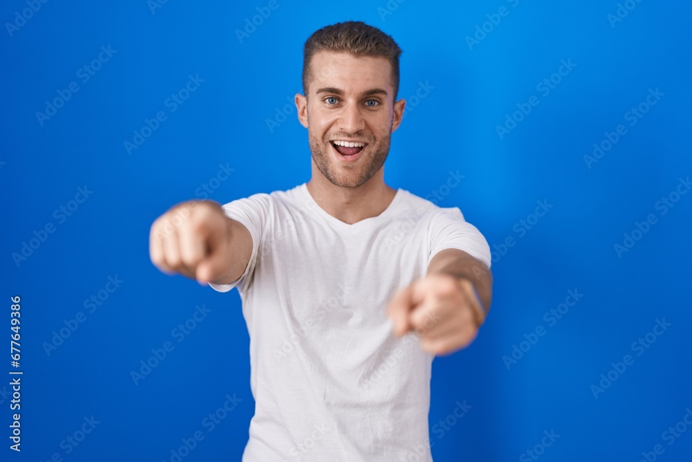 Young caucasian man standing over blue background pointing to you and the camera with fingers, smiling positive and cheerful Young caucasian man standing over blue background pointing to you and the camera with fingers, smiling positive and cheerful