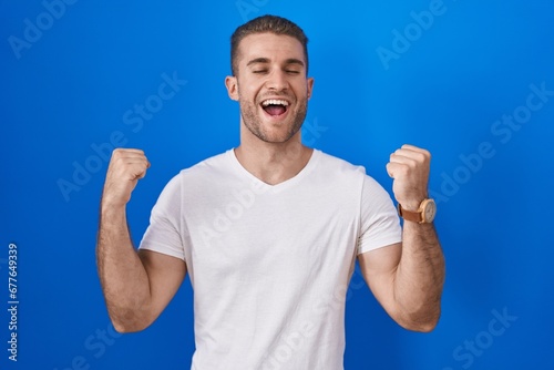 Young caucasian man standing over blue background celebrating surprised and amazed for success with arms raised and eyes closed. winner concept.