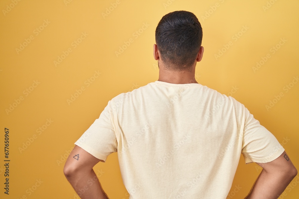 Handsome hispanic man standing over yellow background standing backwards looking away with arms on body Handsome hispanic man standing over yellow background standing backwards looking away with arms on body