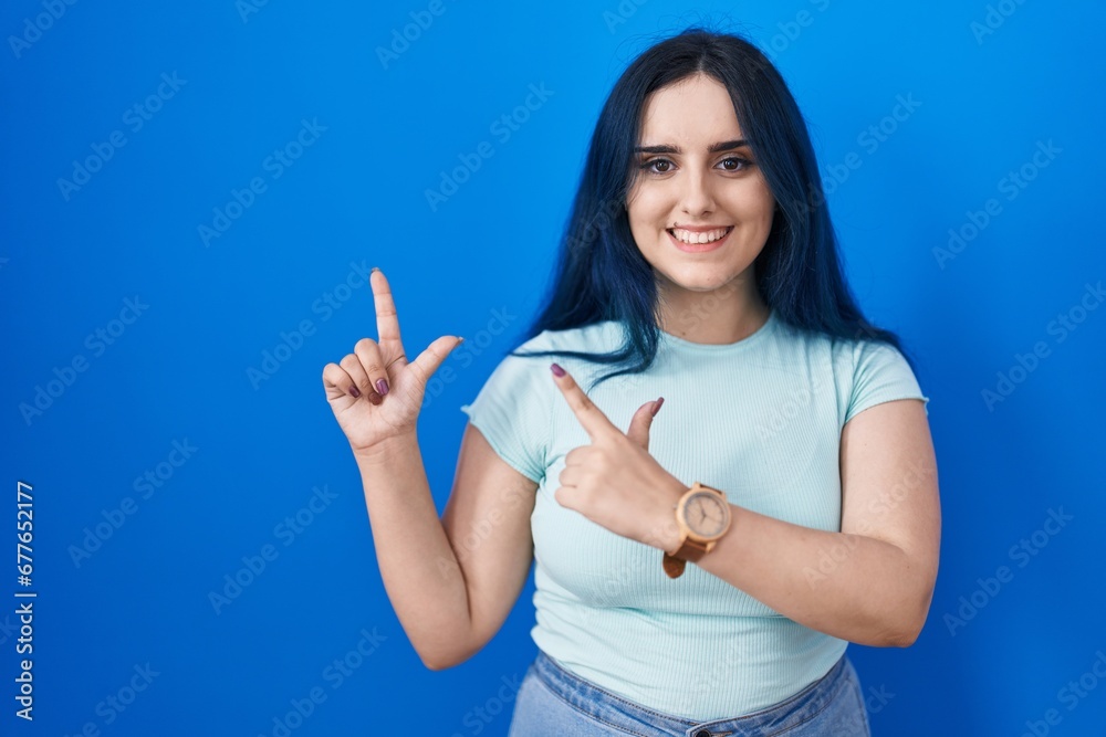 Fototapeta premium Young modern girl with blue hair standing over blue background smiling and looking at the camera pointing with two hands and fingers to the side.