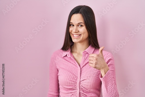 Young hispanic woman standing over pink background doing happy thumbs up gesture with hand. approving expression looking at the camera showing success.