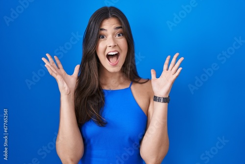 Hispanic woman standing over blue background celebrating mad and crazy for success with arms raised and closed eyes screaming excited. winner concept