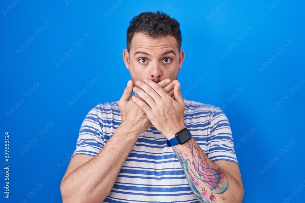 Young hispanic man standing over blue background shocked covering mouth with hands for mistake. secret concept. Young hispanic man standing over blue background shocked covering mouth with hands for mistake. secret concept.