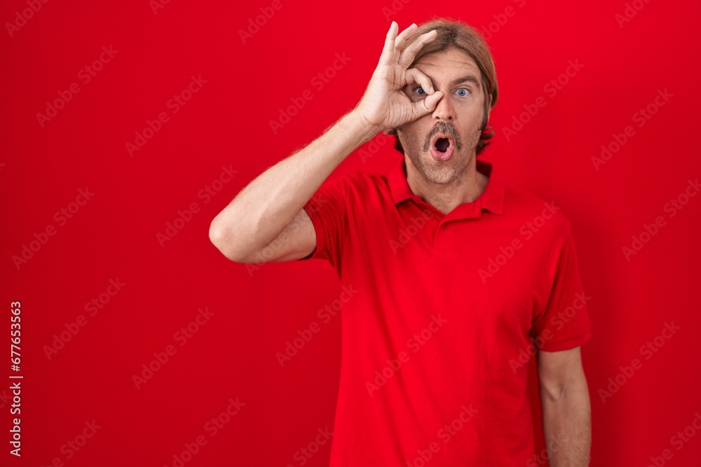 Caucasian man with mustache standing over red background doing ok gesture shocked with surprised face, eye looking through fingers. unbelieving expression. Caucasian man with mustache standing over red background doing ok gesture shocked with surprised face, eye looking through fingers. unbelieving expression.