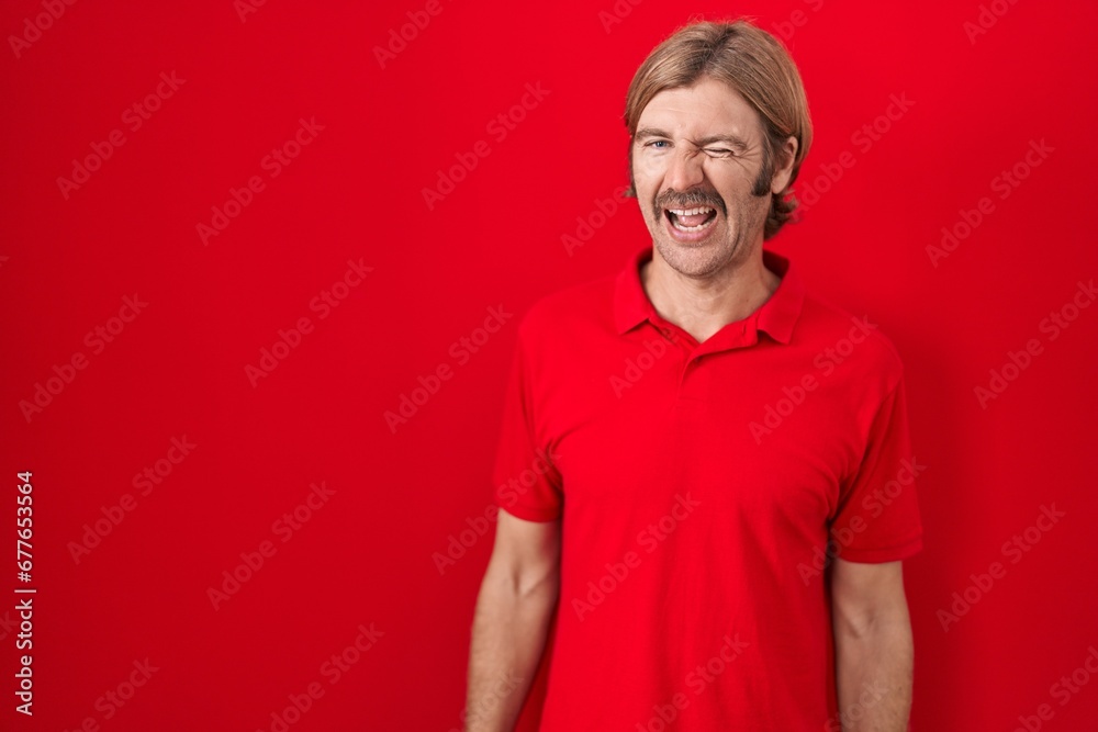 Caucasian man with mustache standing over red background winking looking at the camera with sexy expression, cheerful and happy face. Caucasian man with mustache standing over red background winking looking at the camera with sexy expression, cheerful and happy face.