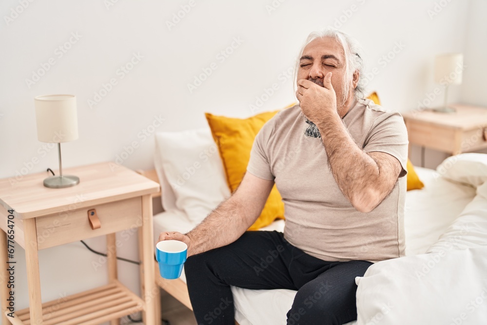 Middle age grey-haired man sitting on bed yawning and drinking coffee at bedroom