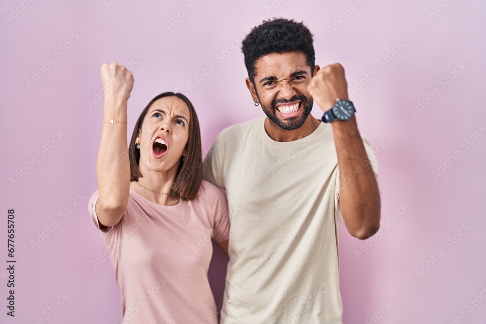 Young hispanic couple together over pink background angry and mad raising fist frustrated and furious while shouting with anger. rage and aggressive concept.