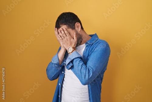Hispanic man standing over yellow background with sad expression covering face with hands while crying. depression concept. © Krakenimages.com