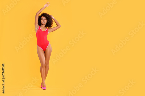 Beautiful woman in bright one-piece summer swimsuit and stylish high heel shoes on yellow background, space for text