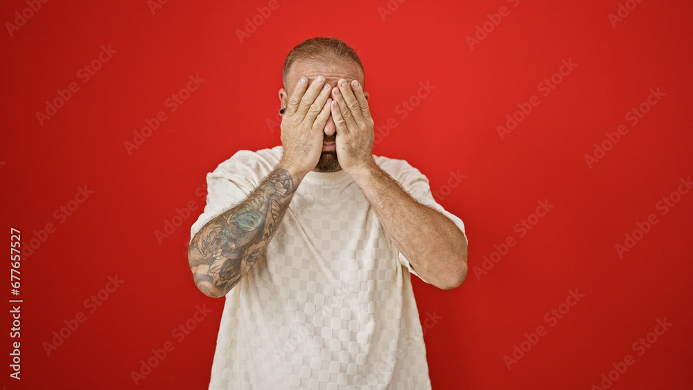 Cool, relaxed young man standing against a red wall, covering eyes, exuding a serious expression. handsome male, casually looking into the camera.