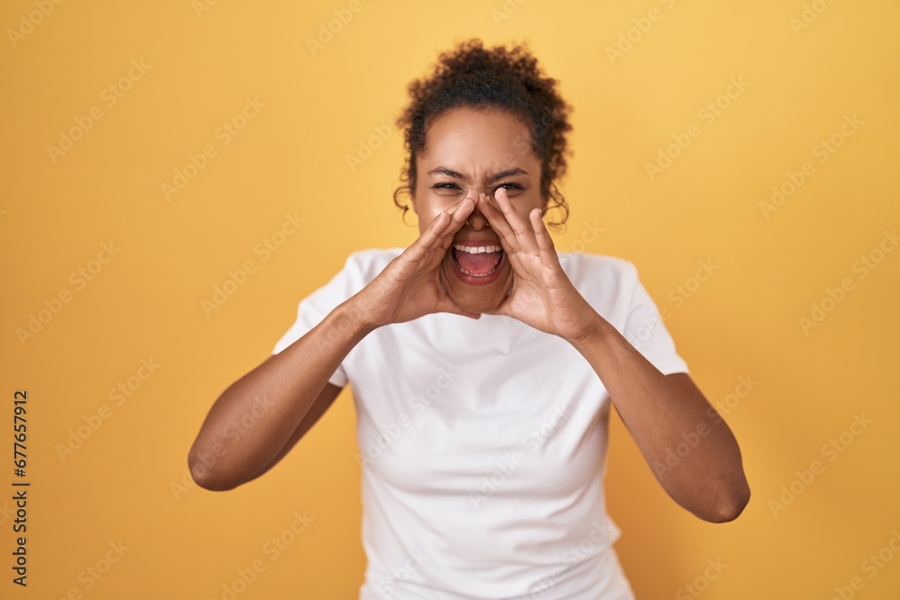 Young hispanic woman with curly hair standing over yellow background shouting angry out loud with hands over mouth Young hispanic woman with curly hair standing over yellow background shouting angry out loud with hands over mouth