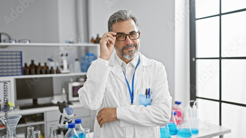 Brimming with confidence, smiling young hispanic man, grey-haired scientist standing in the lab amidst experiments