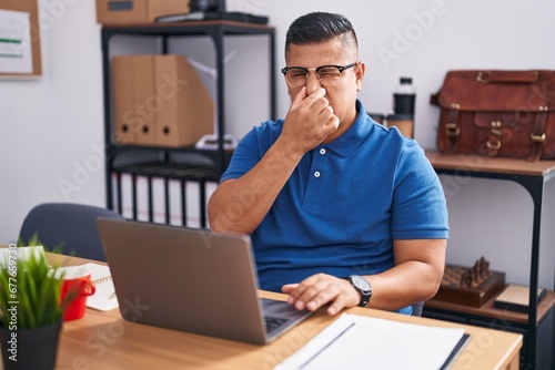 Young hispanic man working at the office with laptop smelling something stinky and disgusting, intolerable smell, holding breath with fingers on nose. bad smell