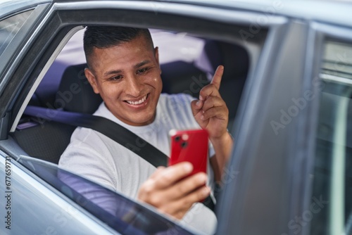 Young hispanic man doing video call with smartphone in the car smiling happy pointing with hand and finger to the side © Krakenimages.com