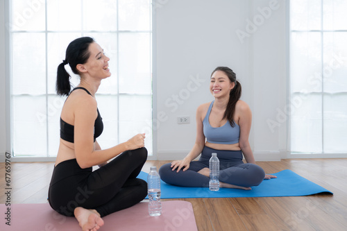Two sporty women sitting rest on yoga mats and drinking water. They are looking at each other and smiling