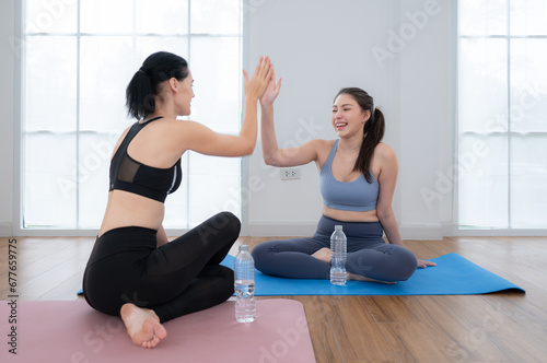 Two sporty women sitting rest on yoga mats and drinking water. They are looking at each other and smiling